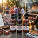 Three bottles of Heath Riles Ultimate Sauce Trio—Sweet, Tangy & Competition BBQ Sauces—rest on a table by the grill, surrounded by food, drinks, and seasonings. People enjoy a perfect BBQ evening under glowing string lights in the background.