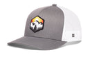The Haka Hat Hexagon Mountain Trees Trucker Hat by Haka Hat is a gray and white snapback with a mesh back, featuring a hexagon patch on the front that showcases a vibrant landscape of trees, sun, and sunset in striking yellow and orange tones—the perfect adventure hat for outdoor enthusiasts.