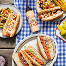 Grilled hot dogs in buns, some topped with diced onions or Terrapin Ridge Farms Harissa Aioli Squeeze. Sides include corn salad, corn on the cob, and pickle spears. A bottle of Terrapin Ridge Farms Harissa Aioli sits on a blue checkered tablecloth.