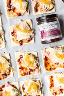 Square dough pieces topped with shredded cheese, ham, and a spicy bacon jam are arranged on parchment paper. A jar of Terrapin Ridge Farms Terrapin Ridge Hot Pepper Berry Bacon Jam is placed among them.