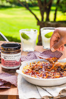 A hand dips a blue corn chip into a cheesy baked dip topped with Terrapin Ridge Farms Hot Pepper Berry Bacon Jam. Nearby are a jar of the savory jam, extra chips, and two cocktails garnished with lime on an outdoor table.