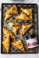 Golden-brown pastry triangles are arranged on a patterned baking tray, accompanied by Terrapin Ridge Farms Hot Pepper Berry Bacon Jam—the perfect savory jam for any cheese board.