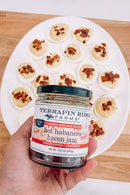 A hand holds a jar of Terrapin Ridge Farms Terrapin Ridge Hot Habanero Bacon Jam in front of deviled eggs topped with bacon—ideal as a sweet and spicy spread or charcuterie board topping on a wooden surface.