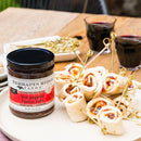 Terrapin Ridge Farms Hot Pepper Bacon Jam sits beside rolled tortilla appetizers, perfect for charcuterie boards. Two glasses of red wine and a small dish are in the background on a wooden table.