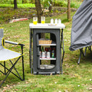 The DirectDeals Folding Camping Storage Cabinet with 3 Shelves and Carry Bag stands on grass next to a foldable chair and tent, holding drinks, glasses, a basket on top, and bottles and plates inside.