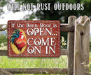 The If The Barn Door Is Open, Come On In - Metal Sign by Lone Star Art hangs on a wooden gate with a rooster design, set against green grass and trees. Will not rust outdoors.