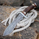 The Texan Knives Damascus Steel Western Bowie Knife – 12" features a patterned blade, Damascus steel guard, stag horn handle, and comes with a leather sheath. It rests on a coiled rope atop rough-cut logs.