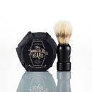 A black-handled shaving brush stands beside a wrapped package labeled “The Immaculate Beard Shave Gift Set” by The Immaculate Beard on a reflective white surface—an ideal gift for anyone who values a classic hot shave and premium natural shave soap.