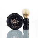 A Shave Soap Puck by The Immaculate Beard, wrapped in black paper, sits next to a shaving brush with a black handle—both reflected on a white surface—making this set an ideal choice for those with sensitive skin.