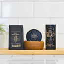 Three premium grooming essentials from The Immaculate Beard—beard wash, Conditioning Natural Beard Balm in a wooden bowl, and beard oil—are artfully arranged on a wooden tray against a white tiled background, with a blurred plant in the corner. This sophisticated display showcases The Immaculate Beard’s natural care products, designed for effortless styling and nourishing of your beard.