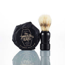A round, wrapped Dark Shave Soap Puck from The Immaculate Beard and a shaving brush with a black handle and light bristles stand side by side on a reflective white surface.