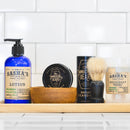 A wooden tray holds lotion, deodorant, a shaving brush, a wooden bowl, and The Immaculate Beard Conditioning Natural Beard Balm—all featuring vintage-style Immaculate Beard branding—set against a white tiled background. This curated collection spotlights natural beard care essentials designed for elevated grooming and effortless beard styling.