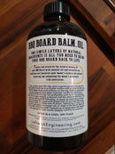 A dark bottle labeled BBQ Board Balm® Care Pair Wax and Oil from BBQ Boards® rests on wood. The label notes it conditions and restores BBQ boards, lists coconut oil and essential oil among ingredients, and includes care directions.
