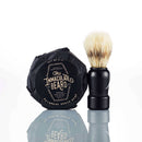 A round bar of The Immaculate Beard Immaculate Beard Dark Shave Soap Protective Lather Sensitive Men in black wrapping sits beside a shaving brush with a black handle, both reflected on a white surface.
