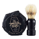 The Immaculate Dense Shave Soap For Smooth Men Wet Shaving by The Immaculate Beard, presented in round black packaging, sits beside a black-handled shaving brush with white bristles—both reflected on a white surface. Ideal for traditional wet shaving and gentle enough for sensitive skin.