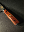 A close-up of the Spaceman Knives Interstellar Kiritsuke Knife by Spaceman Knives, featuring a polished wooden handle with decorative metal rivets and a patterned Damascus blade, resting on a dark surface.