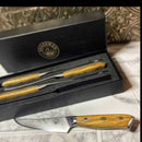 A close-up of a Spaceman Knives Interstellar Series steak knife with a patterned blade and olive wood handle rests on marble, while a black box behind it holds two matching knives from this refined set, all crafted by Spaceman Knives.
