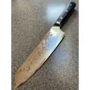 The Spaceman Knives Interstellar Kiritsuke Knife by Spaceman Knives features a patterned Damascus AUS-10 steel blade and a black handle, shown up close as it rests on a speckled gray countertop.