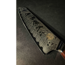 A close-up of the Spaceman Knives Interstellar Kiritsuke Knife blade, featuring a distinctive wavy Damascus pattern and circular logo, crafted from AUS-10 steel by Spaceman Knives, set against a dark background.