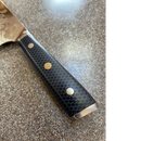 A close-up of the Spaceman Knives Interstellar Kiritsuke Knife handle shows a black hexagonal honeycomb pattern with three metal rivets on a speckled countertop. The blade displays an eye-catching Damascus finish.