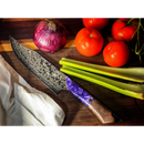 The Spaceman Knives Gravity Series Chef Knife, featuring a patterned AUS-10 steel blade and a purple-brown handle, rests on a wooden cutting board beside an onion, vine tomatoes, celery stalks, and a white towel.