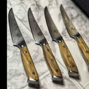 Four Spaceman Knives Interstellar Series steak knives by Spaceman Knives, featuring patterned steel blades and elegant olive wood handles, are neatly arranged in a row on a marble countertop.