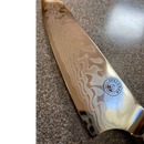 A close-up of the Spaceman Knives Interstellar Kiritsuke Knife, featuring a wavy Damascus-finish blade and Space Man logo on the handle, resting on a speckled countertop.