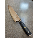 The Spaceman Knives Interstellar Kiritsuke Knife, featuring a patterned Damascus blade and black textured handle, is displayed on a speckled kitchen countertop.