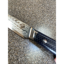 A close-up of the Spaceman Knives Interstellar Utility Knife, featuring a patterned AUS-10 steel blade and a dark honeycomb handle, resting on a speckled countertop.
