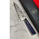 The Spaceman Knives Interstellar Petty Chef Knife, featuring a patterned Damascus blade and blue textured handle, rests on marble beside its black box with gold emblem, highlighting the elegance of Spaceman Knives.