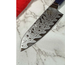 A close-up of the Spaceman Knives Interstellar Petty Chef Knife by Spaceman Knives, featuring a patterned blade, rests on a white marble surface next to a folded red cloth.