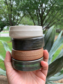 A hand holds a stack of four Shave Bowls by The Immaculate Beard—ideal for whipping up a rich, moisturizing lather with your favorite shave soap puck. The elegant white, black, brown, and green glazes of each ceramic bowl stand out beautifully against the softly blurred trees and plants in the background.
