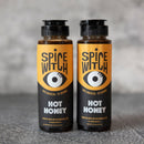 Two bottles of Spice Witch Hot Honey Duo by Spice Witch, with bold yellow and black labels featuring a stylized eye, stand side by side on a gray surface against a blurred gray background.
