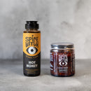 The Spice Witch Hot Honey & Chili Crisp Bundle from Asheville, NC, features hot honey and chili crisp black garlic, both with an illustrated eye logo, displayed on a gray surface.