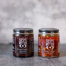 Two glass jars from Spice Witch's Chili Crisp Box of 2 sit side by side on a gray surface, labeled “Black Garlic + Chili Crisp” and “Chili Crisp.”.