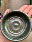 A hand holds The Immaculate Beard Shave Bowl, a small, handmade ceramic bowl with a glossy green and black layered glaze and a textured circular pattern inside—ideal for creating a rich lather from your favorite shave soap puck. The blurred background accentuates the bowl’s striking finish.