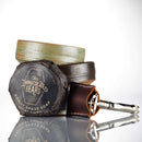 A bar of The Immaculate Beard shave soap, wrapped and labeled, rests next to two stacked ceramic bowls and a metal razor in a brown leather case—all included in the Immaculate Premium Wet Shaving Kit For Men With Ceramic Bowl And Soap—arranged elegantly on a white surface with a bright background.