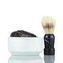 The Immaculate Beard Shave Gift Set sits against a white background, featuring a black-handled shaving brush alongside a white ceramic bowl containing a wrapped natural shave soap bar, capturing the essence of a traditional hot shave.