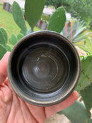 A hand holds The Immaculate Beard Shave Bowl—a small, dark, handmade piece—outdoors, with green cactus pads and other plants softly blurred in the background.