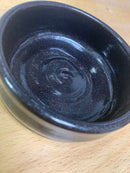A small, empty, glossy black ceramic bowl from The Immaculate Beard sits on a light wooden surface, its smooth interior reflecting light—a perfect handmade Shave Bowl for creating a rich, moisturizing lather with your favorite shave soap puck.