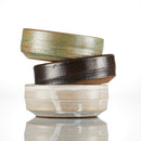 Three round ceramic Shave Bowls by The Immaculate Beard, shown in green, dark brown, and off-white, are stacked unevenly on a reflective white surface. Each bowl features a rustic, handmade look with subtle glaze variations—ideal for building a rich, moisturizing lather with your favorite shave soap puck.