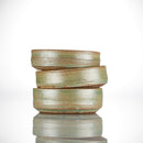 Three greenish-brown ceramic Shave Bowls from The Immaculate Beard are stacked unevenly on a reflective surface against a white background. Featuring a handmade, rustic look with horizontal textured lines, these bowls are perfect for creating a rich, moisturizing lather when paired with your favorite shave soap puck.