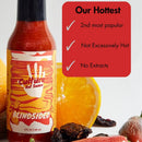 A bottle of Culture Hot Sauce Blindsided by Culture Hot Sauce—our bold Carolina Reaper sauce—rests beside colorful citrus fruits and a variety of peppers. A sign displays proudly checked phrases: Our Hottest, 2nd Most Popular, Not Excessively Hot, and No Extracts.