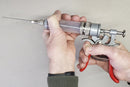 Someone holds the SpitJack MAGNUM Meat Injector—Complete Kit with Deluxe Case and Four Needles—featuring a long needle and red trigger, ideal for BBQ competitors, against a plain background.