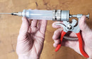 Someone holds The SpitJack MAGNUM Meat Injector—by SpitJack—with a clear barrel, metal needle, and red trigger; this complete kit includes four needles and a deluxe case, shown against a wooden background.