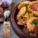 A wooden bowl of shrimp and sausage jambalaya, a hearty and gluten free dish, sits next to garlic cloves on burlap. In the foreground is a container of Hickman's BBQ Cajun Gluten Free Keto Friendly Natural BBQ Rub 4 oz by Hickman's BBQ, highlighting the bold flavors that make this jambalaya truly special.