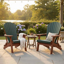 Two Kanyon Living Poly Folding Adirondack Chairs in Dark Green and Cedar Wood sit on a stone patio with green cushions, facing each other across a small table. A white throw and plant rest on the chairs; hydrangeas and trees brighten the sunlit background.