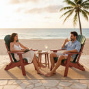 A woman and man relax in Kanyon Living's Kanyon Poly Folding Adirondack Chairs in Dark Green & Cherry Wood on a beachside patio at sunset, sipping drinks with snacks on a small table and an ocean view framed by a swaying palm tree.