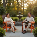 A woman with a cup and a man holding a book sit on Kanyon Living’s Kanyon Poly Folding Adirondack Chairs in Dark Wood and Orange on a garden patio, surrounded by greenery, string lights, and a fountain glowing at sunset.
