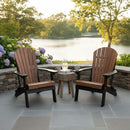 Two Kanyon Living Kanyon Poly Folding Adirondack Chair Deluxe in Antique Mahogany & Black sit on a stone patio by a lake, facing a small round table with drinks and a candle amid hydrangeas and trees in this peaceful outdoor setting.